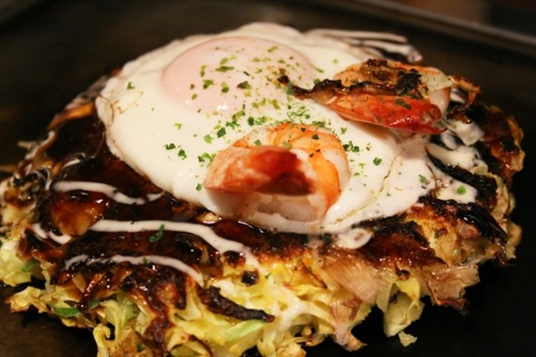 An okonomiyaki on the grill, topped with an egg and shrimp