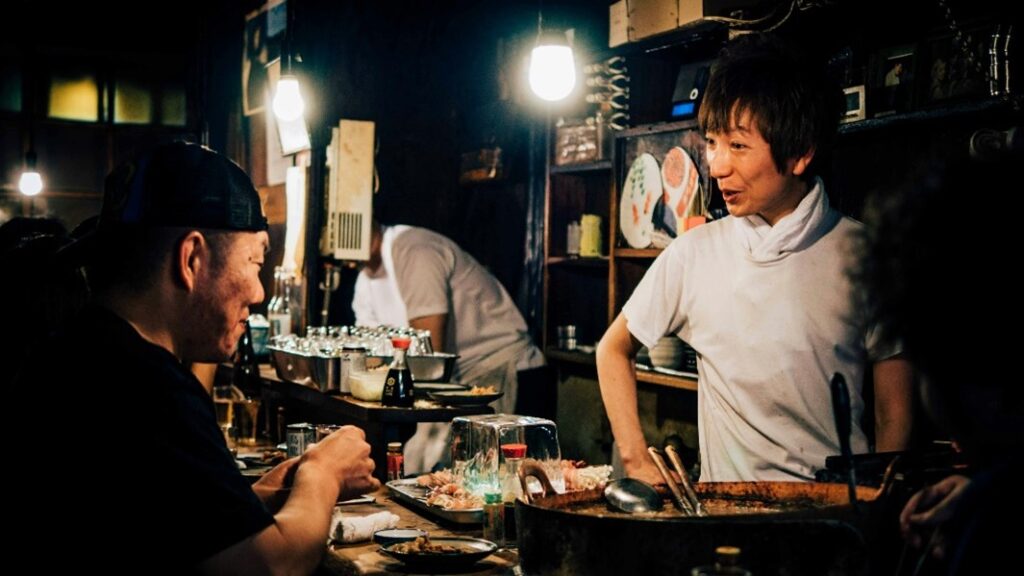 A waiter serving food to a customer at a Tokyo bar while two people engage in conversation.