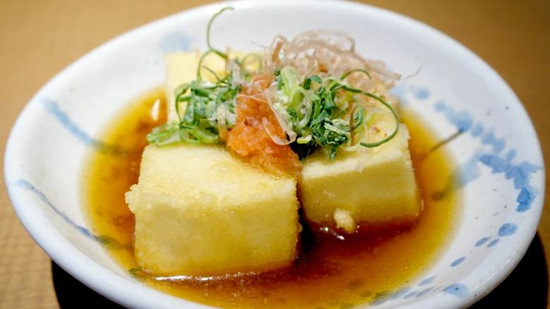 A bowl of agedashi tofu showing three golden-brown fried tofu cubes in a shallow pool of soy-based broth