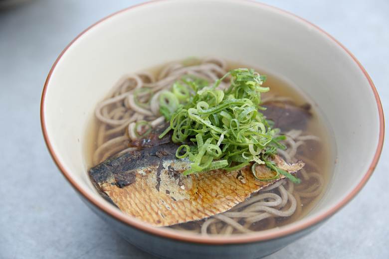Close-up of a bowl of hot soba noodles in clear broth topped with a grilled fish fillet