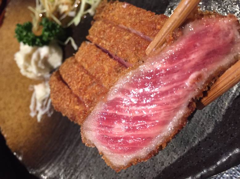 Chopsticks holding a slice of rare-cooked beef katsu with a golden, crispy panko crust, served on a black plate