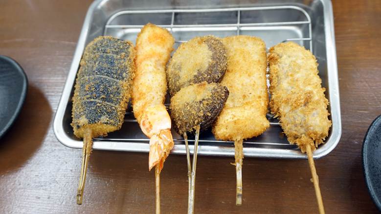 A metal tray with a wire rack holding five assorted kushikatsu skewers