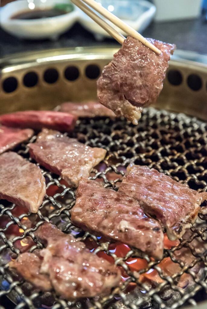 A pair of chopsticks lifting a grilled beef slice from a charcoal yakiniku grill