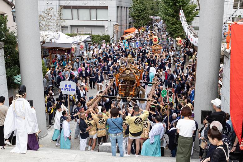 A lively procession at a traditional festival in Tokyo, with people carrying mikoshi (portable shrines), and spectators watching along the street.