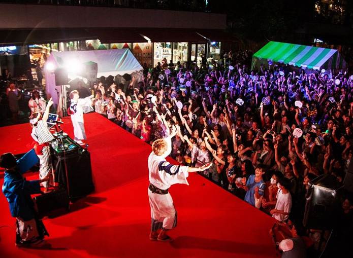 Performers on a brightly lit stage entertaining a lively crowd at a nighttime outdoor event in Tokyo.