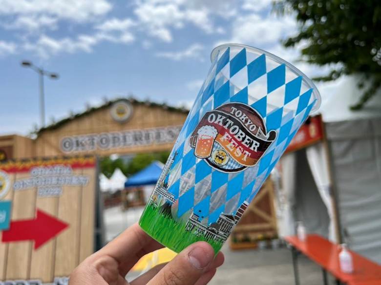 A hand holding a decorative cup from Tokyo Oktoberfest, with the event signage and entrance visible in the background under a partly cloudy sky.