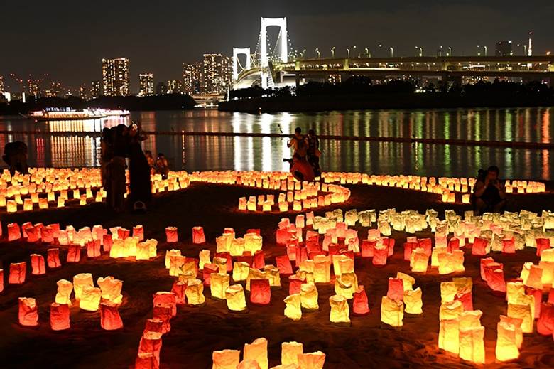 Illuminated paper lanterns arranged beautifully on a beach, with Tokyo's Rainbow Bridge and city skyline glowing in the background at night.