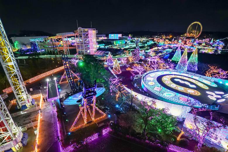 Vibrant, colorful lights illuminating an amusement park in Tokyo at night, showcasing beautifully decorated trees, rides, and a large Ferris wheel in the distance.