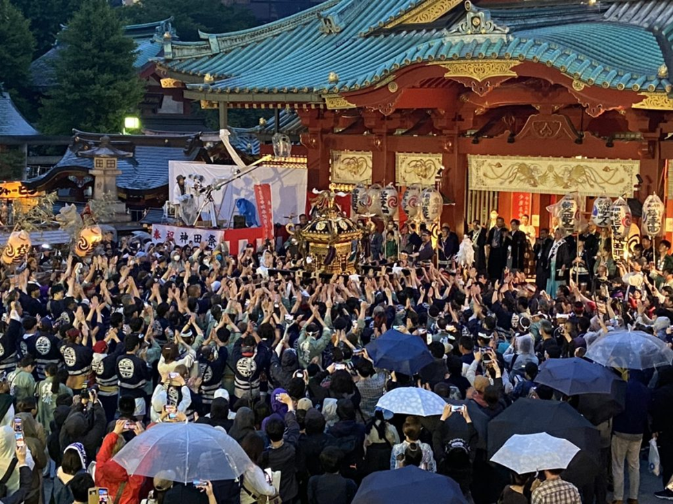 Crowd of festival-goers gathered under umbrellas at dusk in front of a red-and-green‐tiled Shinto shrine