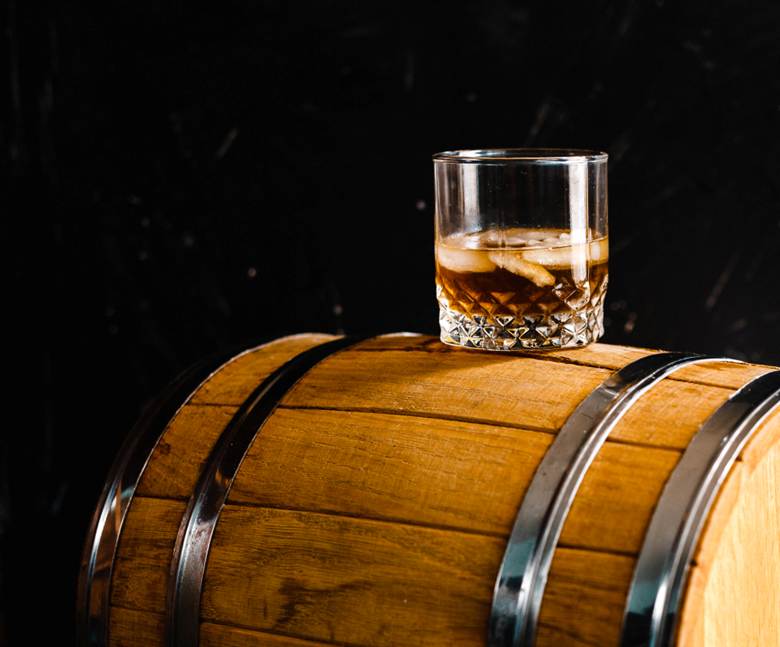 A glass of whisky with ice resting on a wooden barrel, with a dark background.